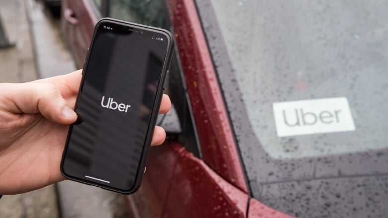 A hand holds an iPhone opened to the Uber app in front of the windshield of a car with an Uber sticker on it.