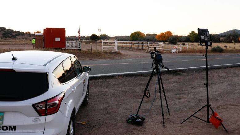 A car and a camera on a tripod are seen on the side of the road outside a fenced property. 