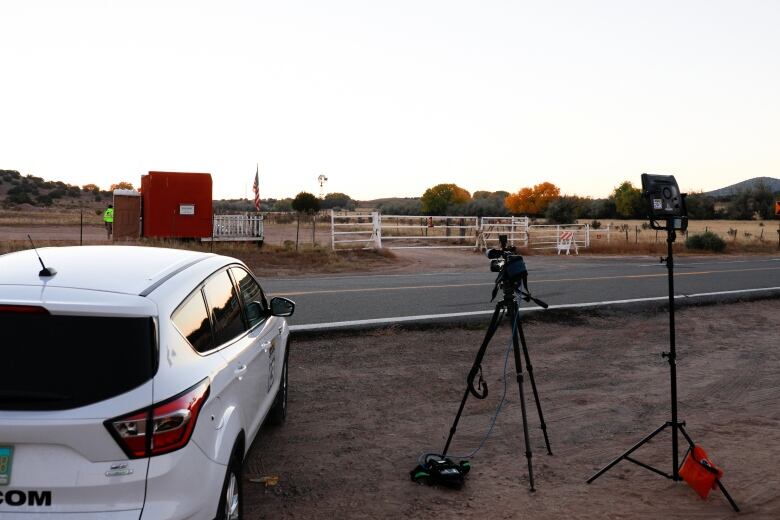 A car and a camera on a tripod are seen on the side of the road outside a fenced property. 