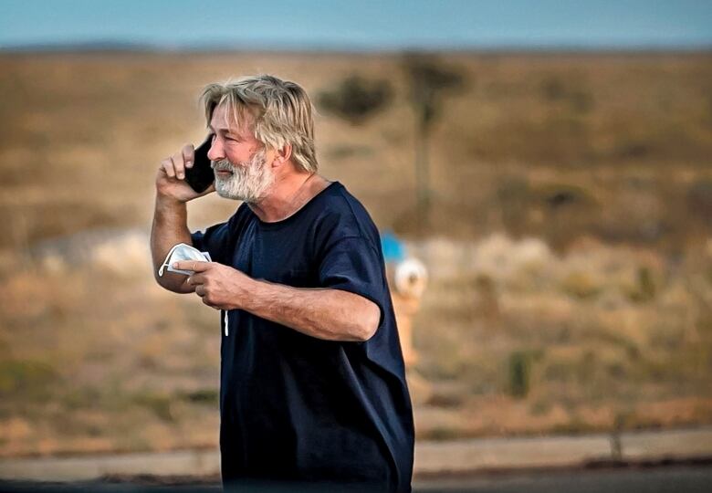 A bearded man standing in a parking lot speaks on a cellphone.