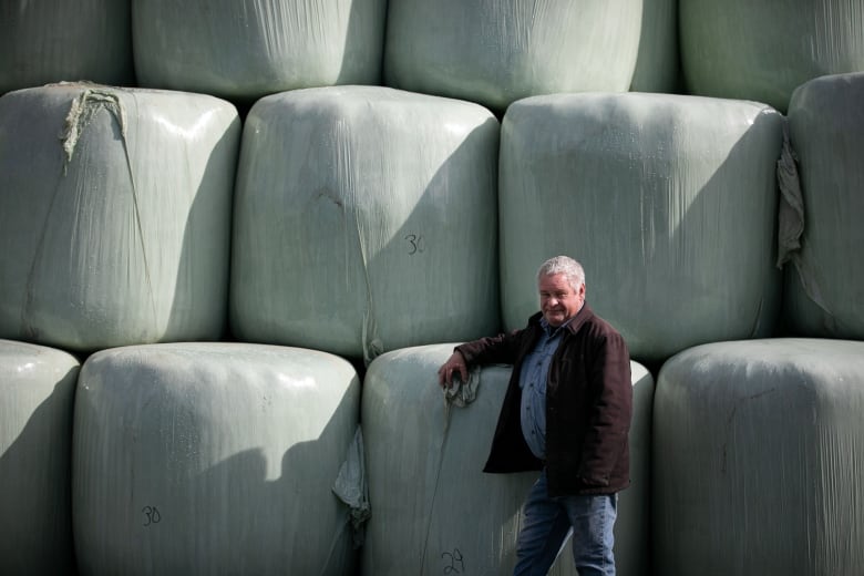 A man in a black jacket and blue shirt is leaning against a wall of stacked crop bales. 