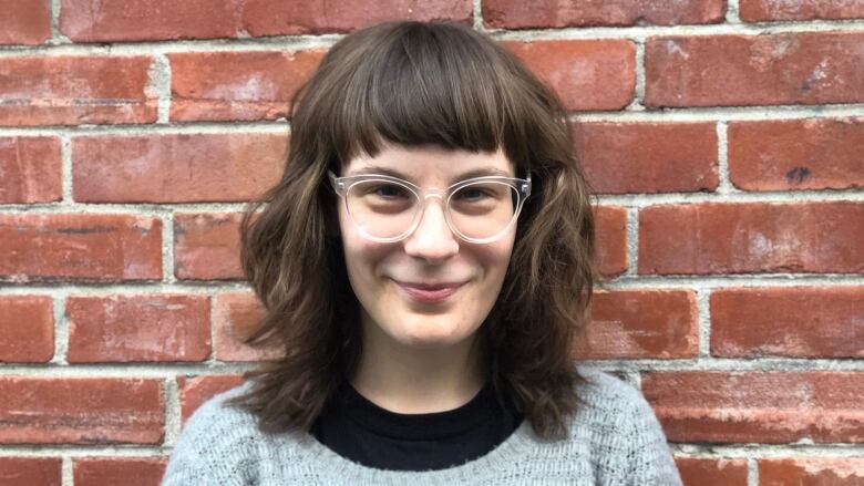 A dark haired woman wearing glasses and a grey shirt smiling and standing in front of a brick wall