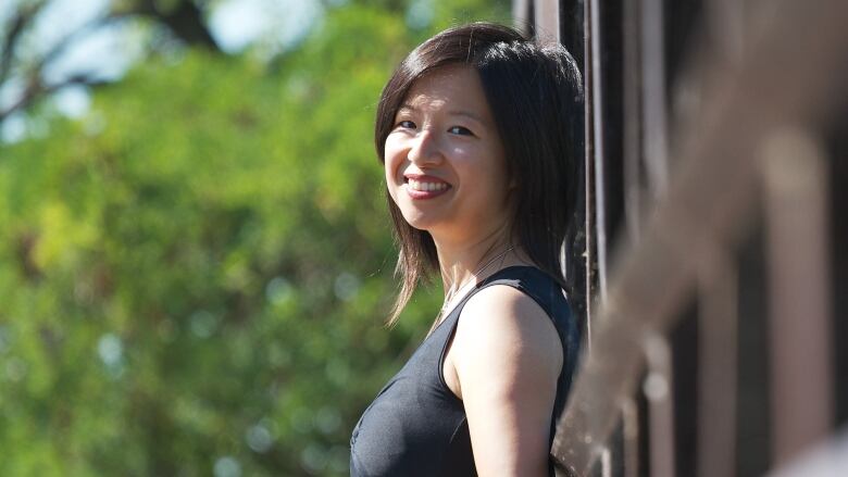 A smiling Asian woman with dark mid-length hair leaning against a wall with some greenery in the background