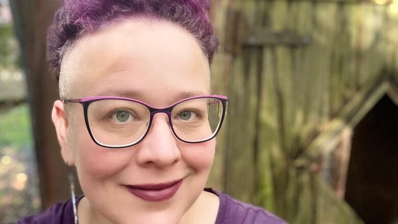 A close-up portrait of a woman with short purple hair, purple rimmed glasses and wearing a purple t-shirt smiling at the camera.