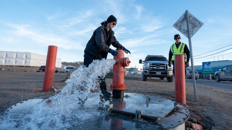 Two people stand by a fire hydrant as water pours out.