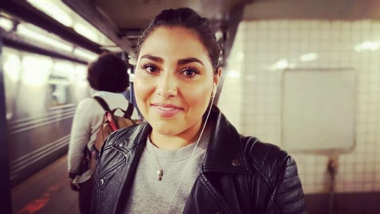 A young woman with her hair pulled back wearing a black leather jacket over a grey sweater and standing on a subway platform