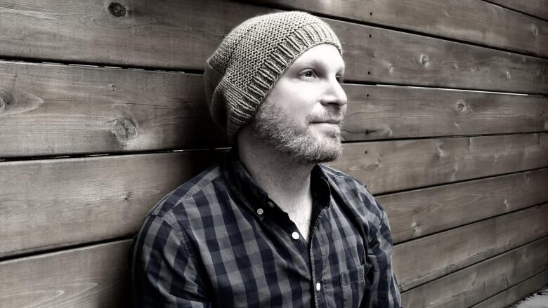 A sepia side portrait of a bearded man wearing a beanie and a plaid shirt standing in front of a wooden slats wall