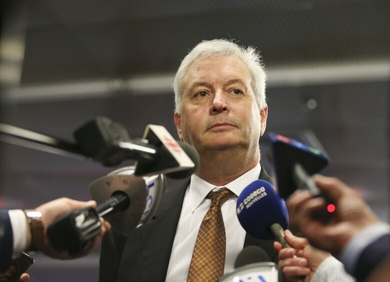 A white man in a suit speaks into several microphones from news agencies.