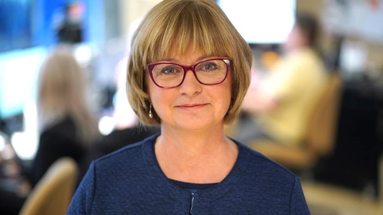 A woman with blonde hair, wearing glasses and a blue dress, looks toward the camera.