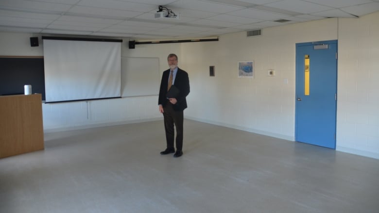 A bearded man wearing a suit stands in an empty classroom 