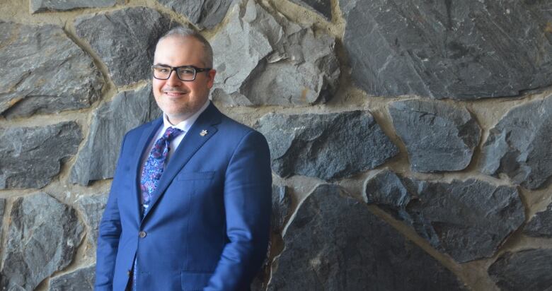 A smiling man in a blue suit stands against a stone wall 