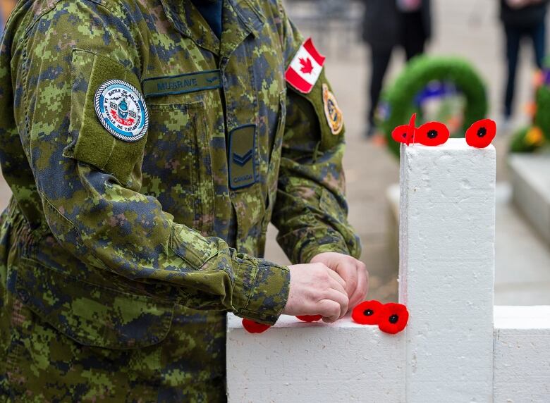 A person in green army fatigues is seen from the mid chest down laying red poppies on a white cross.