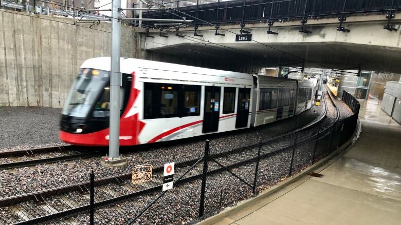 A red-and-white train travels on tracks under a concrete bridge.