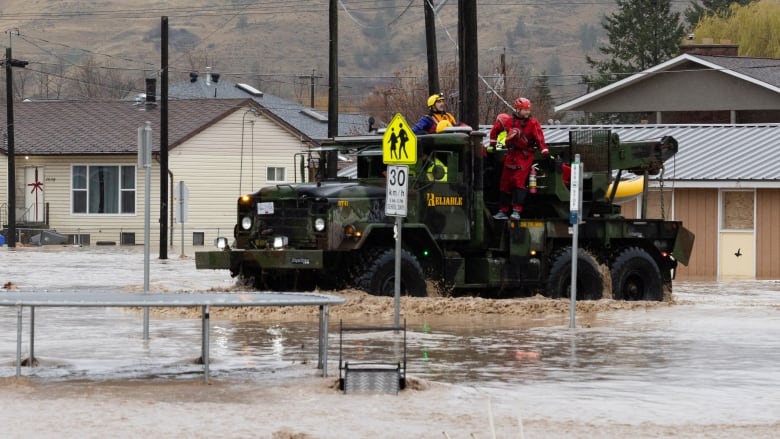 A heavy-duty vehicle drives emergency responders wearing helmets and waterproof suits through a flooded urban street.