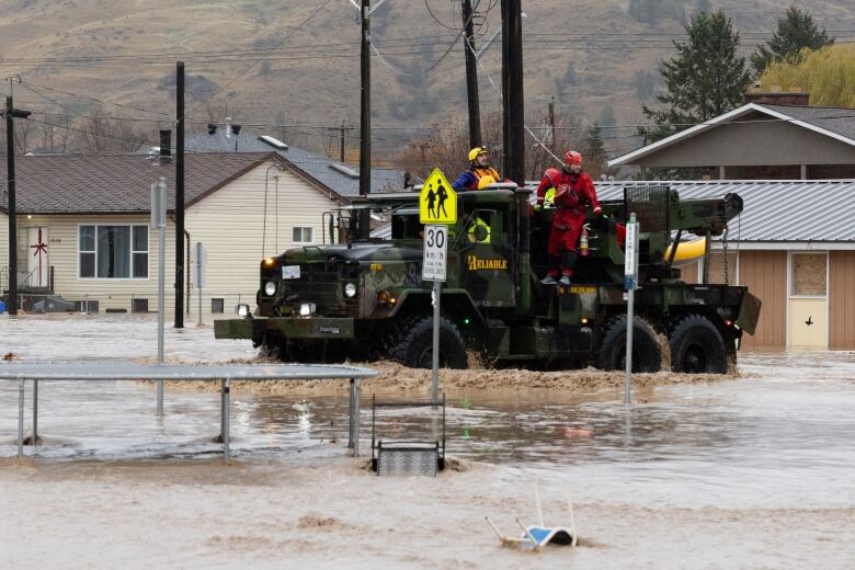 A heavy-duty vehicle drives emergency responders wearing helmets and waterproof suits through a flooded urban street.