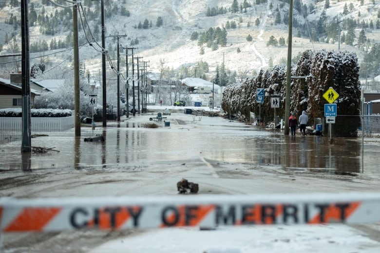 A flooded street is seen behind a barrier, with snow-capped hills visible in the background.