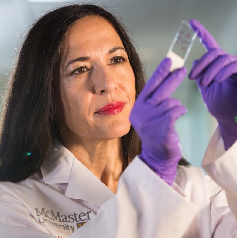 A woman with dark hair wearing a white lab coat and purple lab gloves looks at a medical slide.