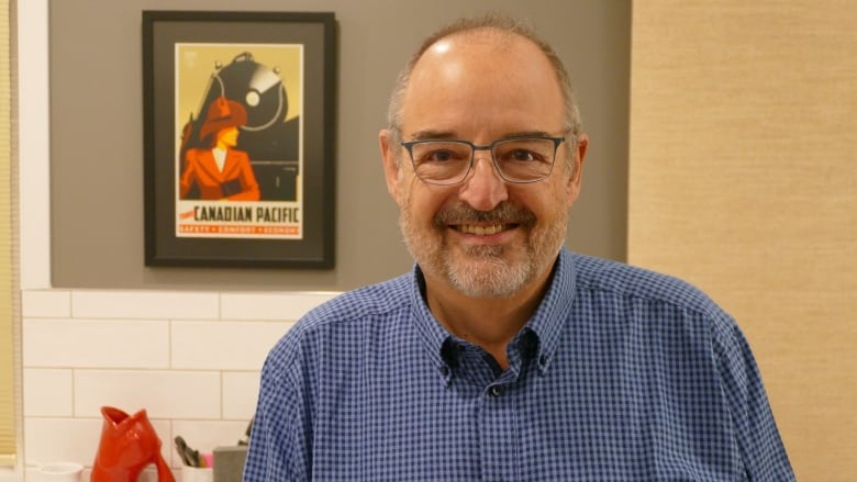 A bald man with a grey and white beard wears a collared shirt and smiles.