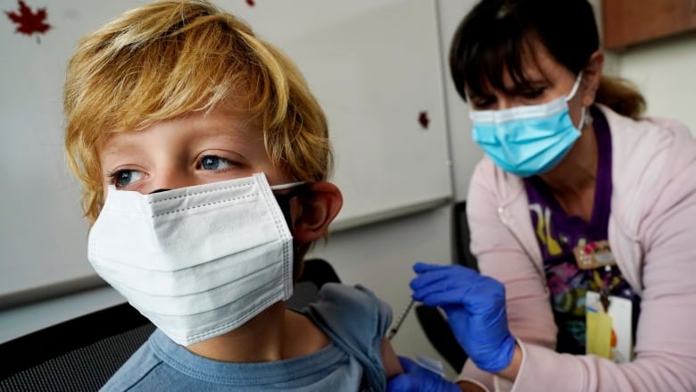 A blond-haired boy wearing a face mask turns his head away from a woman injecting a needle into his upper left arm.