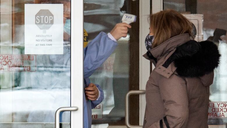 A woman in a mask stands outside the door of a long-term care building as a person wearing protective gear and a mask takes her temperature through the open door.