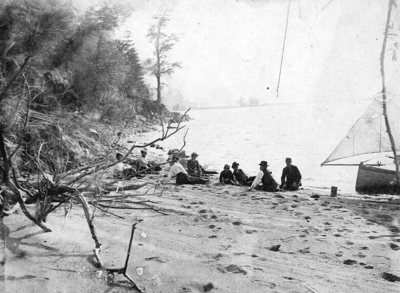 An old black and white of several people in hats and clothing lounging along the shore of a lake with a sailboat to their right.