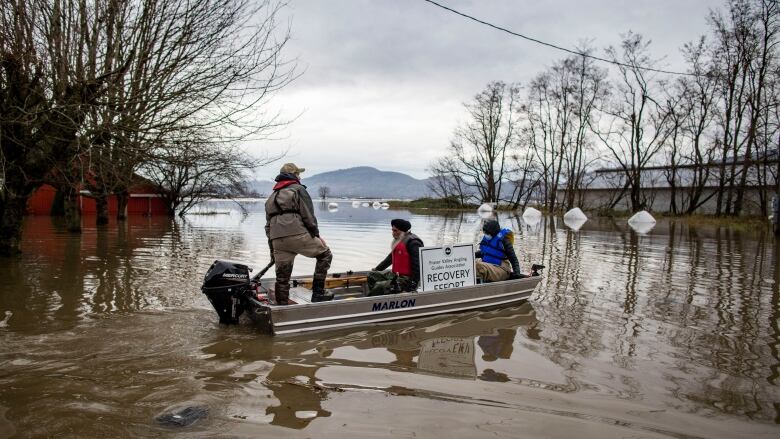 Three people sit in a small tin boat on top of murky brown flood water during the November 2021 floods in B.C.