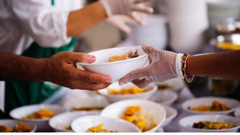 Two hands reach out, from different sides of a table, passing a bowl of food. On the table there are many more bowls of food. 