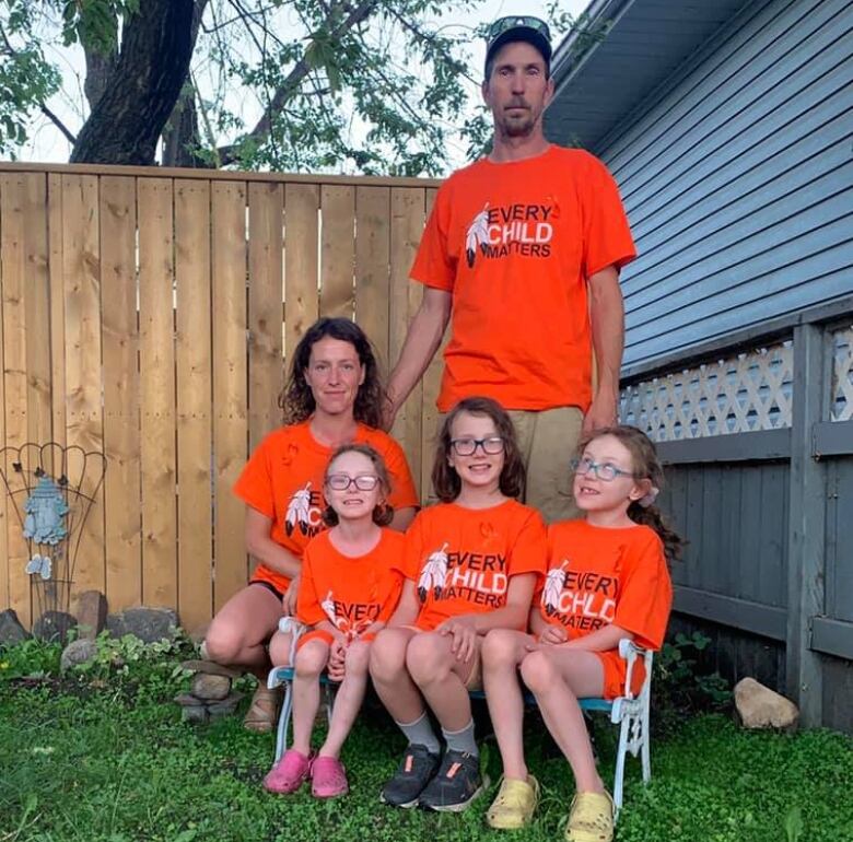 A man stands behind a woman, crouching down, and three young girls sitting in lawn chairs. All wear orange shirts reading 