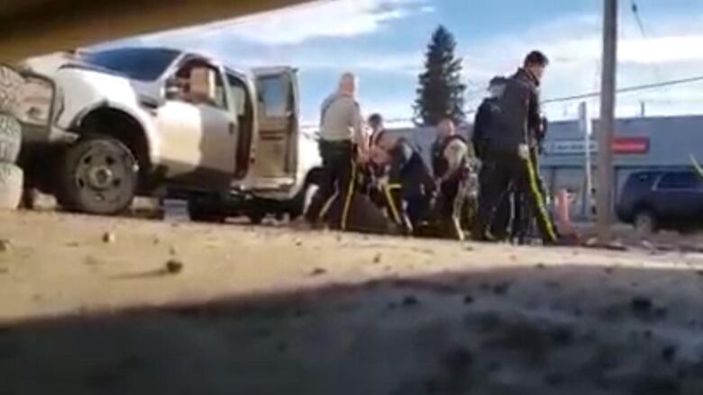 A blurry photo taken from ground level shows numerous RCMP officers in uniform who appear to be struggling with someone, beside a white pick up truck with an open door. 