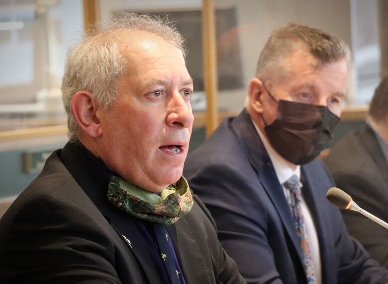 A man in a brown jacket speaks out while another man wearing a blue suit and face mask listens in.