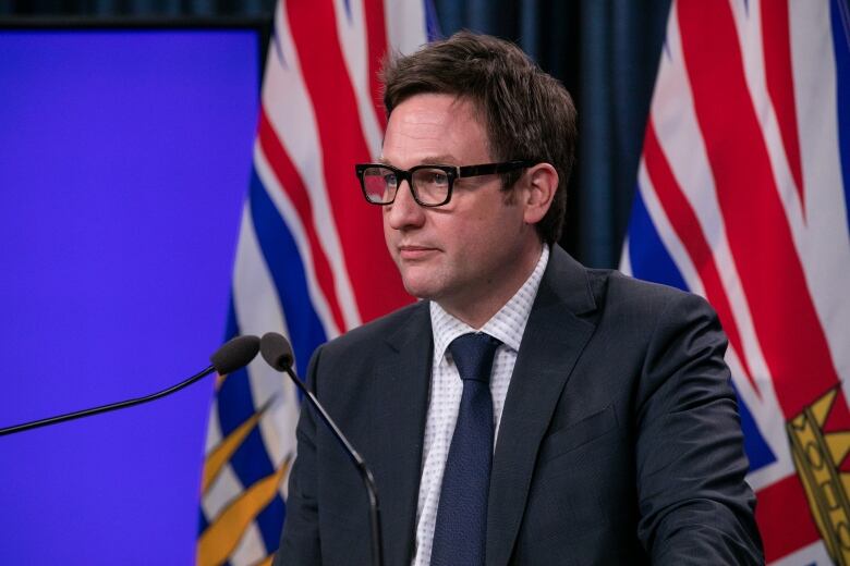 A white man speaks in front of two B.C. flags at a podium.