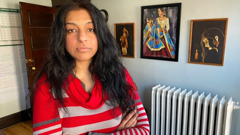 A woman with long black hair, wearing a red and white striped shirt, stands in front of paintings showing women of different ethnicities. 