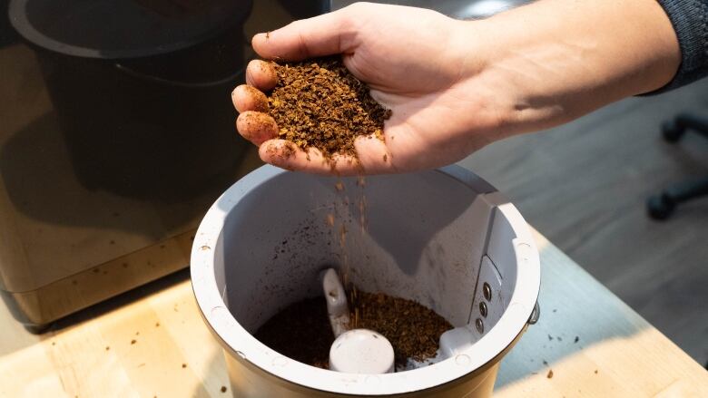 A hand full of a dirt-like substance, being poured into a white cylindrical device. 