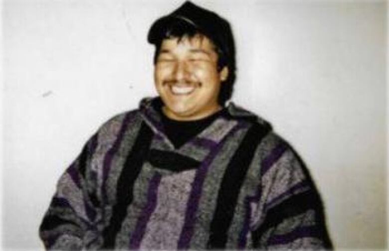 A man wearing a purple, black and grey sweater smiles while leaning against a wall.