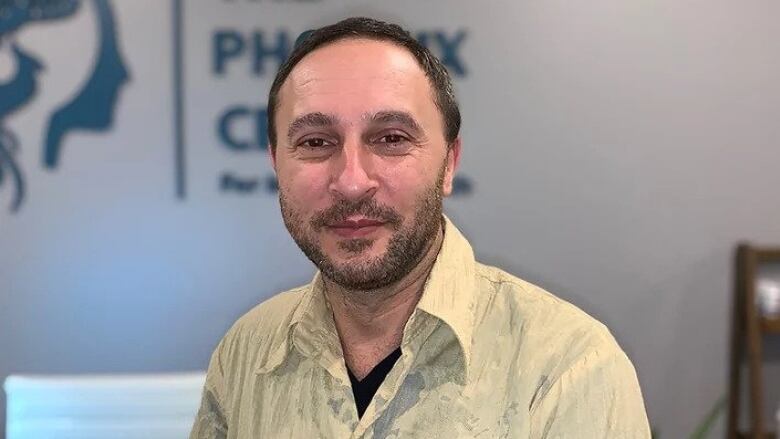 Xander Phoenix looks straight at the camera. He is a white man with black balding hair and a thick stubble, wearing a yellow shirt. A sign behind him reads 'The Phoenix Centre'.