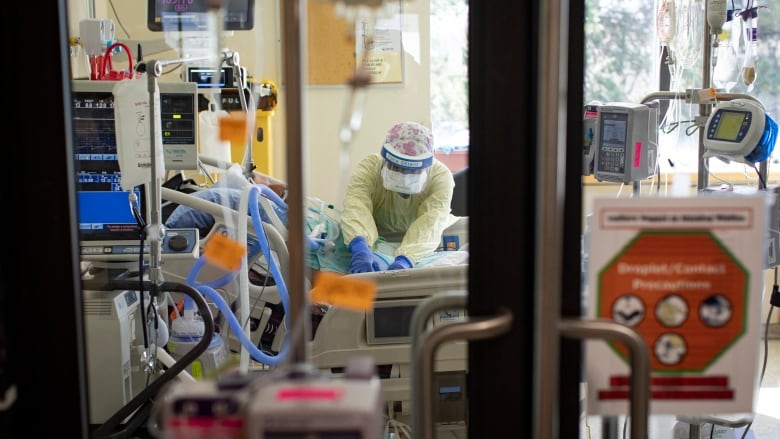 An ICU nurse is seen caring for a patient in a hospital bed through the window of a door.