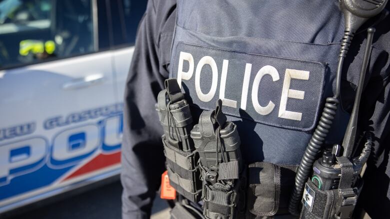 The torso of a police officer wearing a vest that says police. A police car for new Glasgow is in the background.