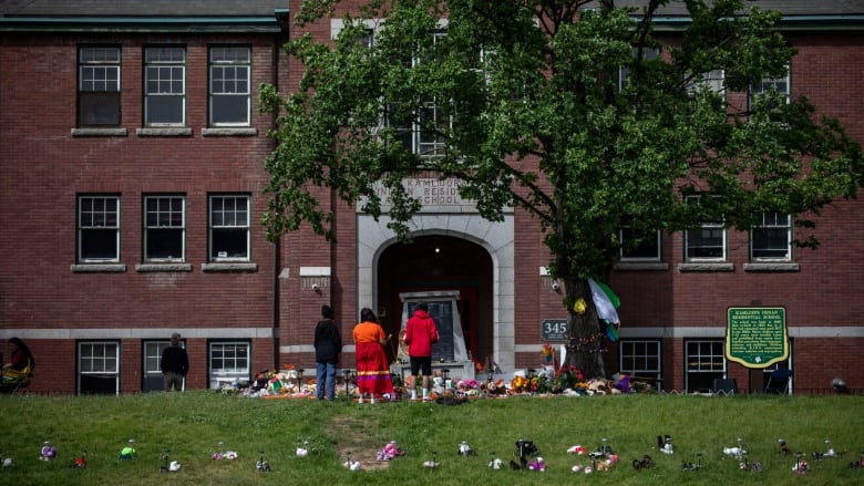 People wearing orange shirts stand in front of a stone monument surrounded by flowers, teddy bears and other tributes in front of the brick building that once housed the Kamloops Indian Residential School.