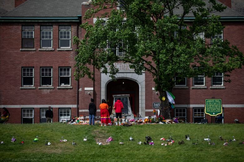 People wearing orange shirts stand in front of a stone monument surrounded by flowers, teddy bears and other tributes in front of the brick building that once housed the Kamloops Indian Residential School.