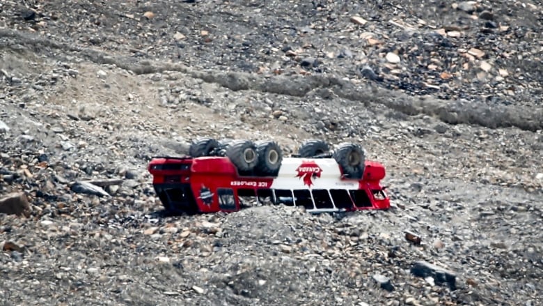 A bus lies on its roof, with wheels in the air, surrounded by rock.