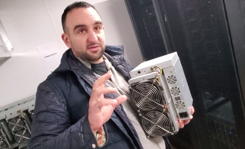 A man in a black jacket holds a piece of equipment that is a small computer known as a miner.