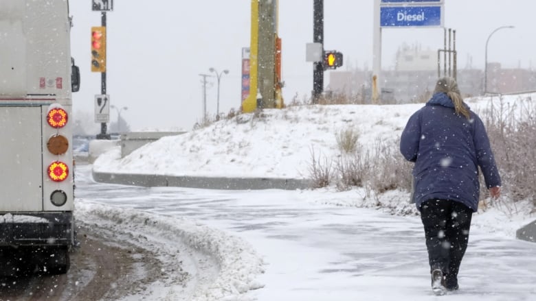 a woman in a blue winter coat walks along a snowy sidewalk as a city bus goes by. 