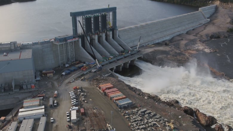 The Muskrat Falls dam seen from overhead while under construction. The dam holds a large body of water in the top left of the photo, while water shoots out into a river at the bottom right. There's construction equipment on a dirt lot at the bottom left.