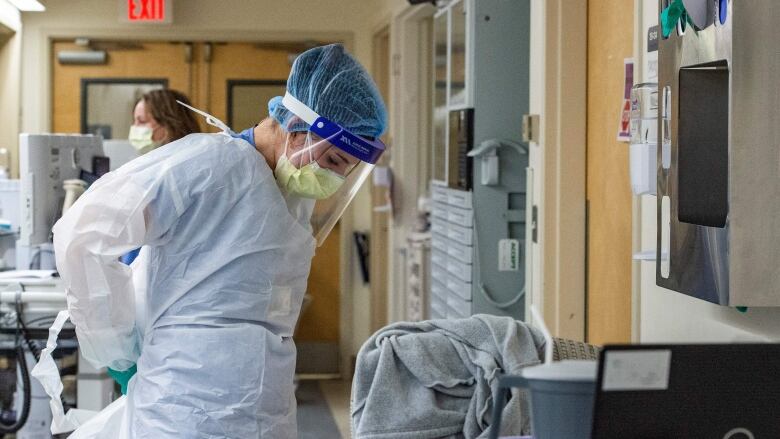 A female health-care worker wearing full personal protective equipment, including a yellow medical mask, face shield and blue latex gloves, does up the ties of her white plastic medical gown behind her back.