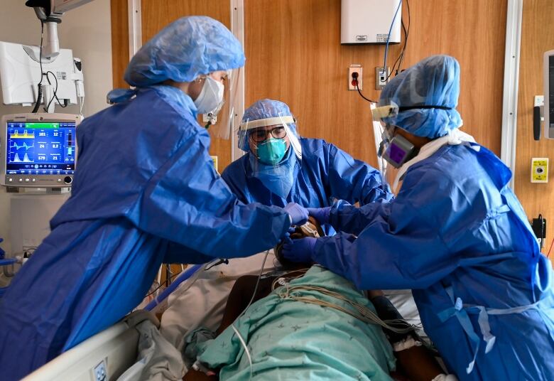 Three hospital staff members in blue gowns and facemasks work on a patient lying on a stretcher.