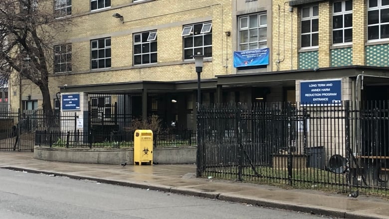 A large building with a needle disposal bin in front of it is shown on a rainy day from the city street