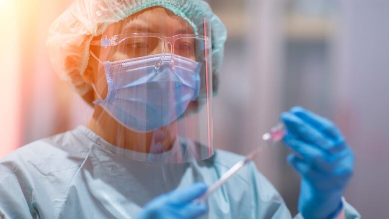 Woman in medical blue scrubs, face shield, mask and gloves holds a syringe.