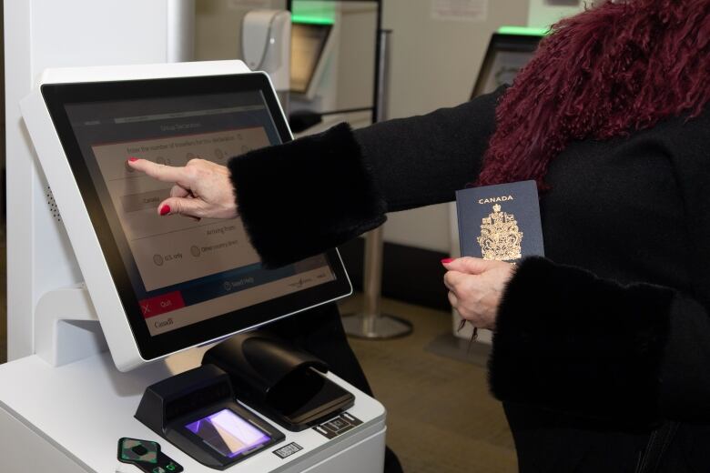 A person presses a touchscreen at an arrival terminal at an airport. They are holding a passport in their other hand.