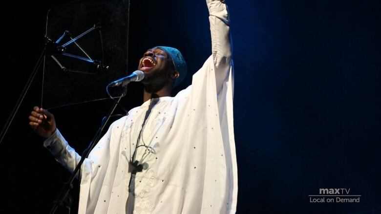 A Black man in a white hat and robe sings into a microphone