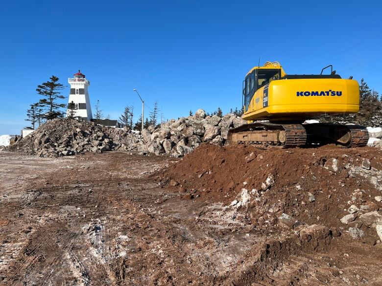 An active construction site with the West Point lighthouse in the background. There is a wall of rocks and a yellow construction vehicle on a mound of dirt. 